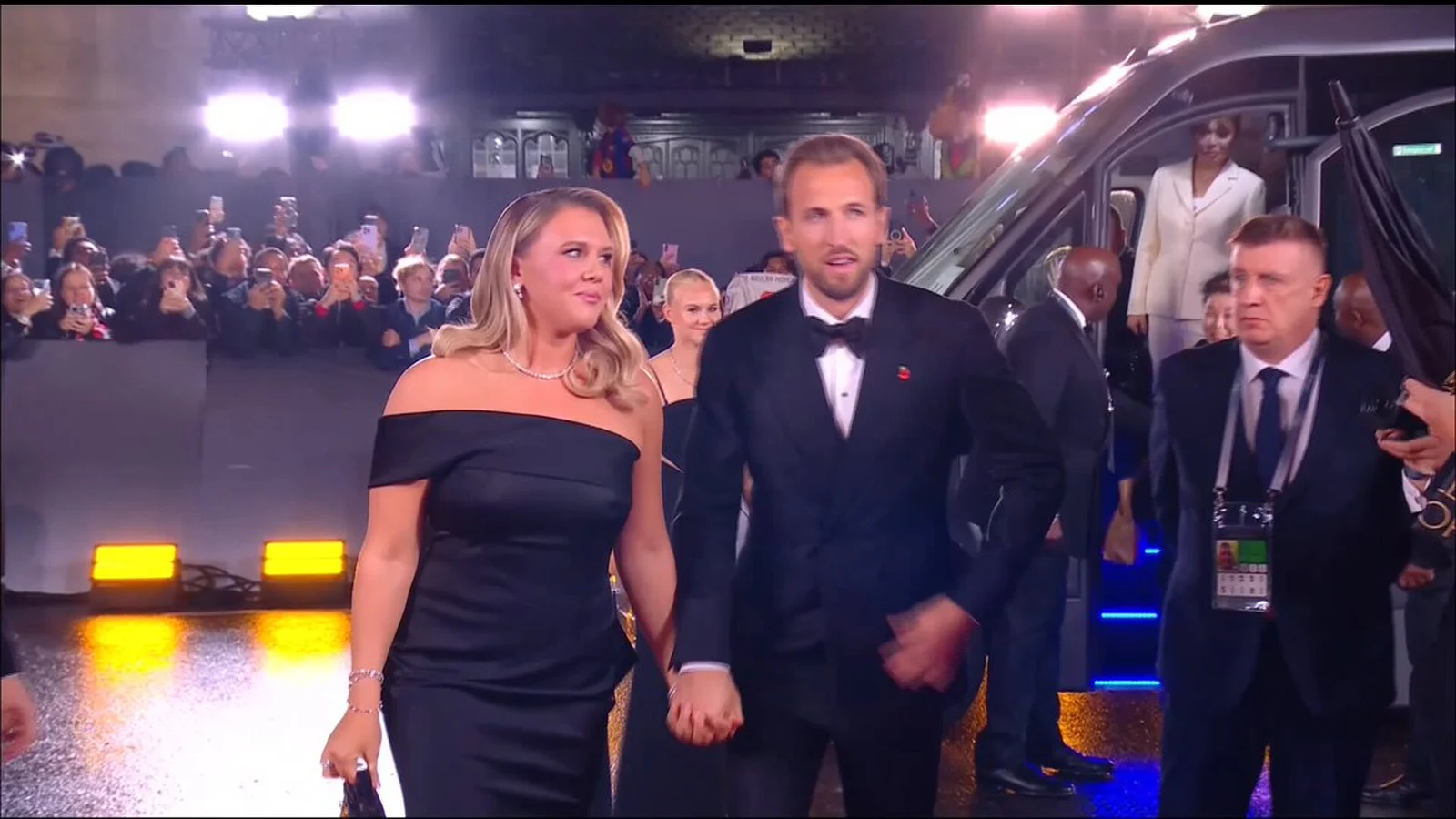 Harry Kane mặc vest đen nắm tay vợ bước trên thảm đỏ trước đám đông giơ điện thoại