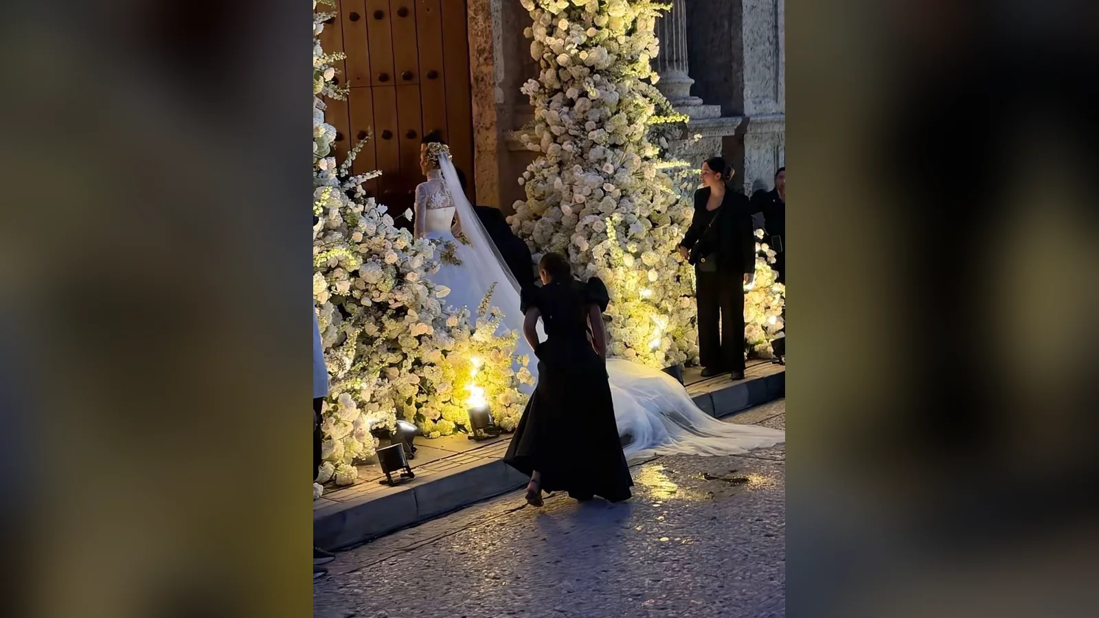 Ariadna Gutiérrez trong váy cưới trắng bước vào cổng nhà thờ phủ đầy hoa trắng, người phụ nữ mặc đồ đen đỡ tà voan dài