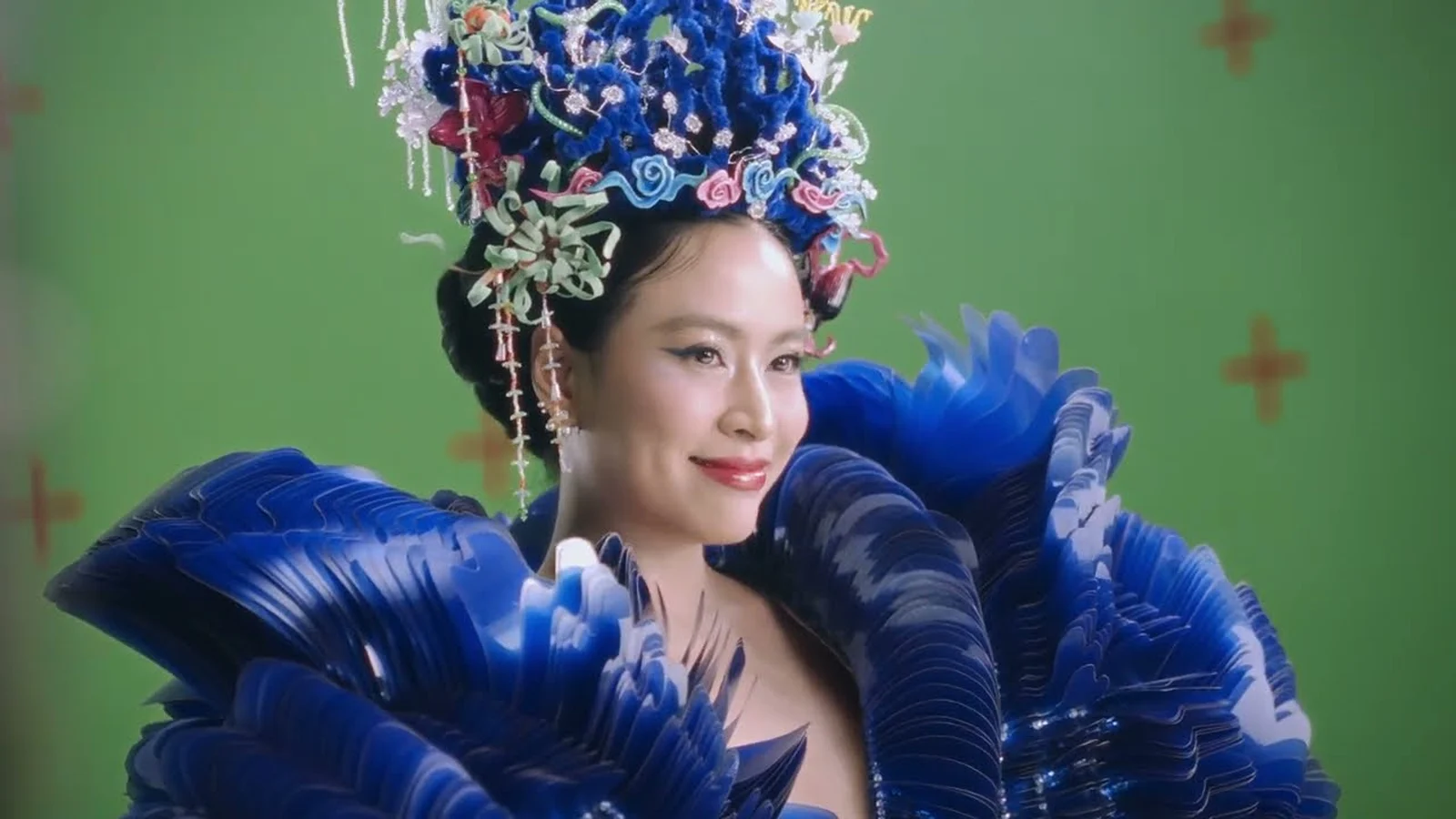 Hoàng Thùy Linh đội mũ kết hoa, mặc áo xanh xếp lớp trước phông xanh ở hậu trường quay MV Thầy Dùi