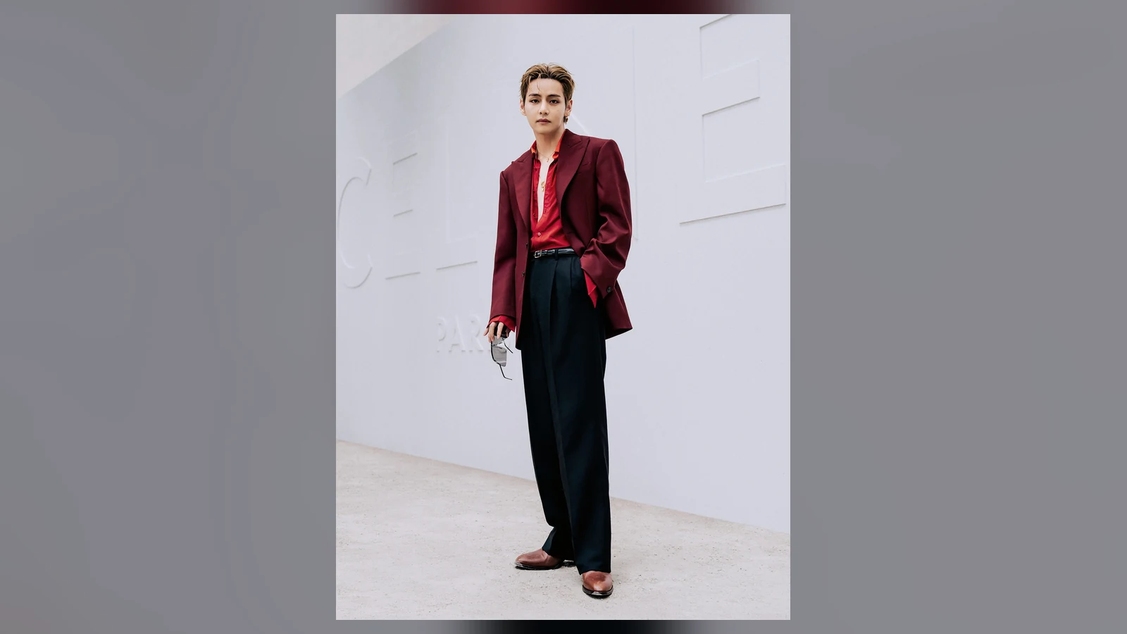 BTS V đứng trước chữ CELINE PARIS, blazer đỏ sẫm, áo đỏ, cầm kính đen