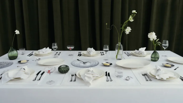 Bàn tiệc bày bộ sưu tập đồ bàn ăn Westwing x Victoria Beckham với đĩa sứ trắng, ly và bình hoa xanh
